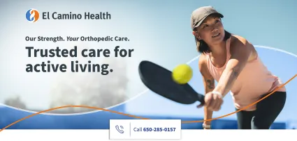 A woman in a pink sleeveless top and gray cap plays pickleball outdoors, reaching forward with a paddle to hit a yellow ball.