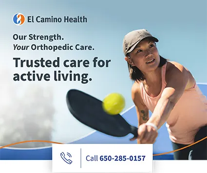 A woman in a pink sleeveless top and gray cap plays pickleball outdoors, reaching forward with a paddle to hit a yellow ball.